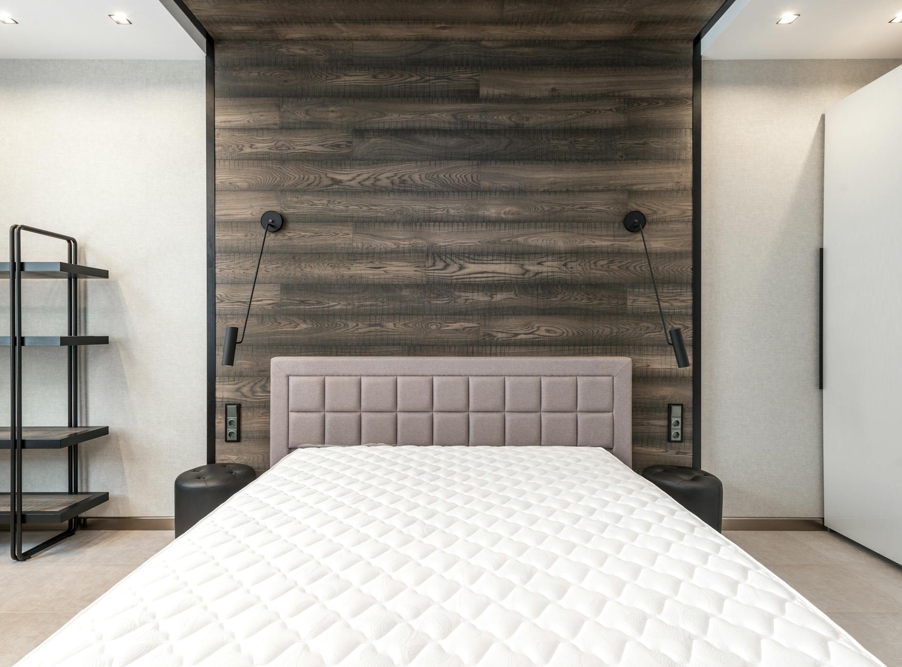 Bao lâu nên giặt nệm một lần? - Comfortable bed with white mattress placed at wall with wooden element in stylish spacious bedroom with lamps and shelves in apartment