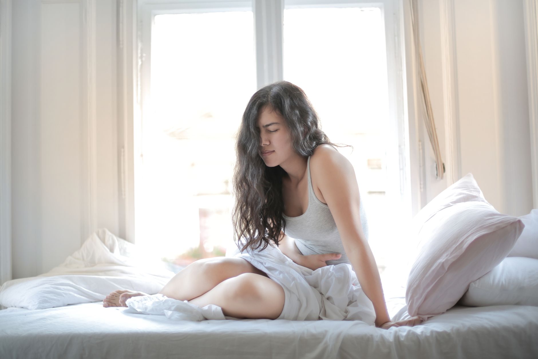 Nệm chống đau lưng cho người ngủ nghiêng - Woman sitting on bed holding her stomach, showing signs of discomfort or pain indoors.