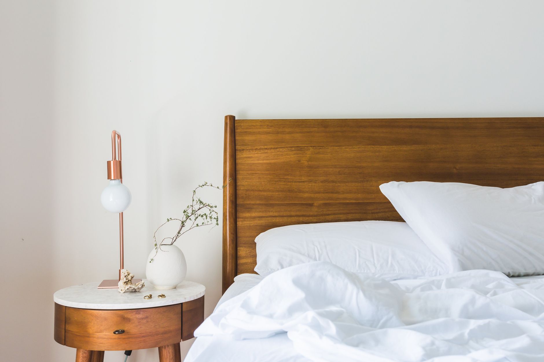 Cách khử mùi hôi trên nệm hiệu quả - A minimalist bedroom features a wooden headboard, white bedding, and a stylish side table lamp.