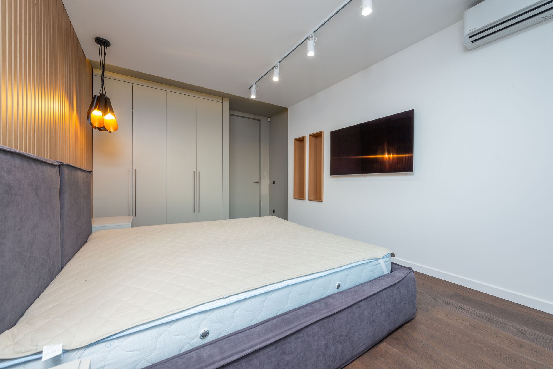 Nằm nệm cao su có bị nóng lưng không - Spacious modern bedroom featuring a gray bed and wall-mounted TV with ambient lighting.