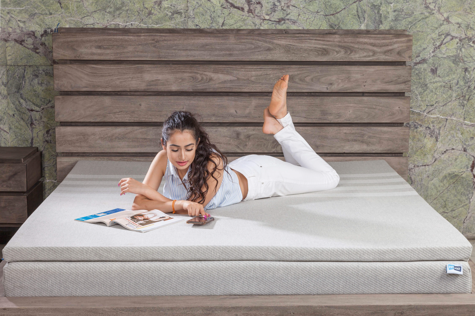 Cách dùng topper nệm để tăng độ êm. - A young woman relaxing on a bed with a magazine and cellphone, set against a stylish wooden headboard.