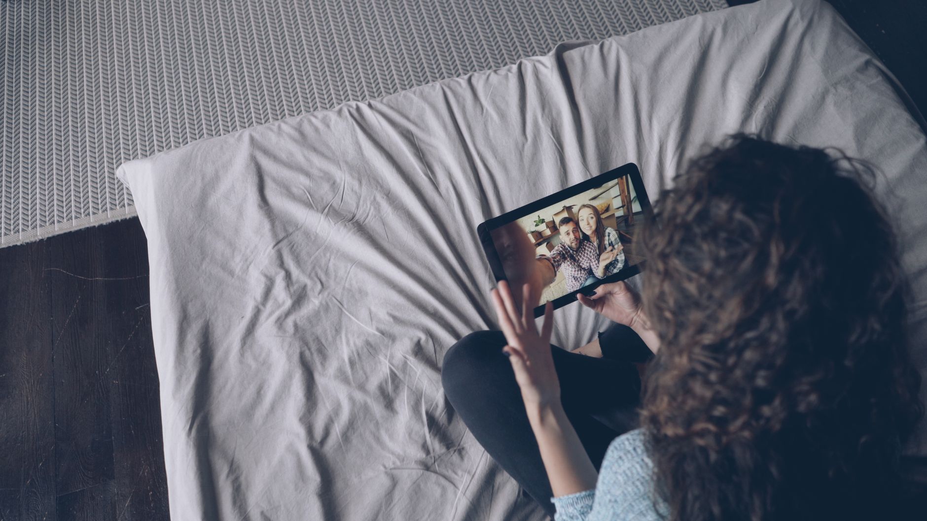 Cách diệt rệp giường trên nệm - A woman uses a tablet for a video call with friends while sitting on a bed indoors.