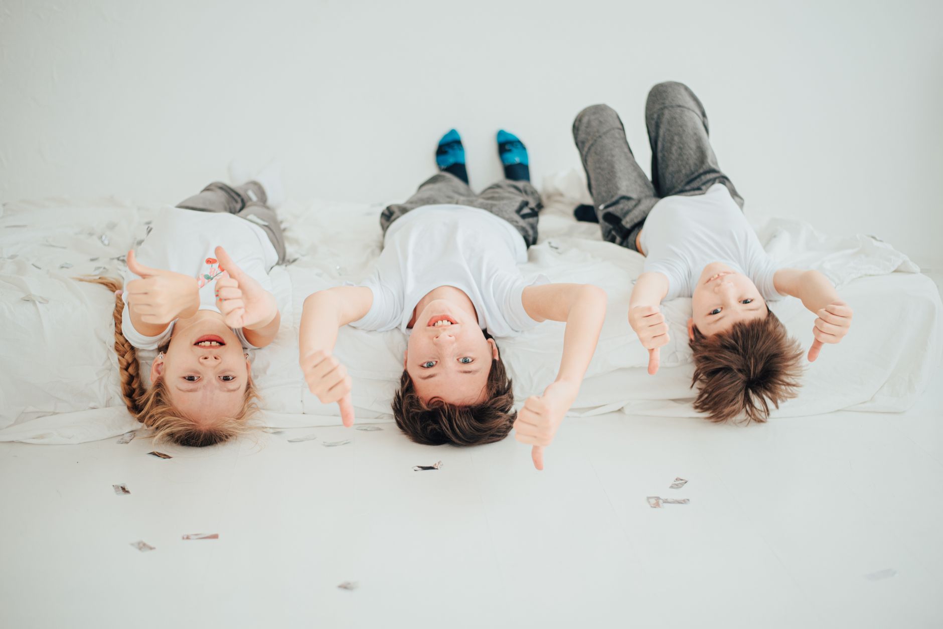 Trẻ em có nên nằm nệm memory foam - Three kids lying upside down on a bed, smiling and giving thumbs up in a bright studio.