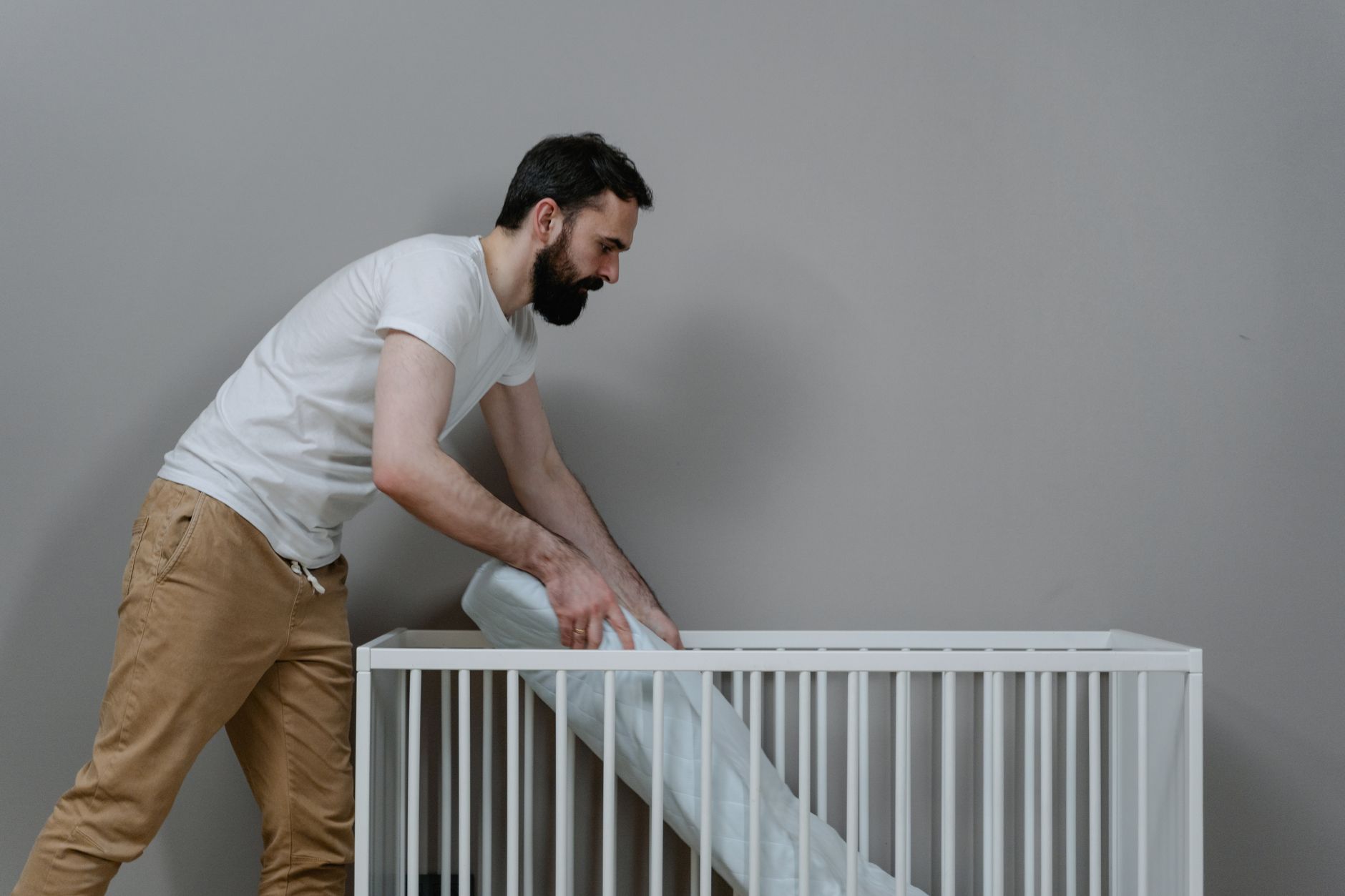 Nệm cho bé từ 1 đến 3 tuổi loại nào tốt - A bearded man setting up a crib in a minimalistic nursery room.