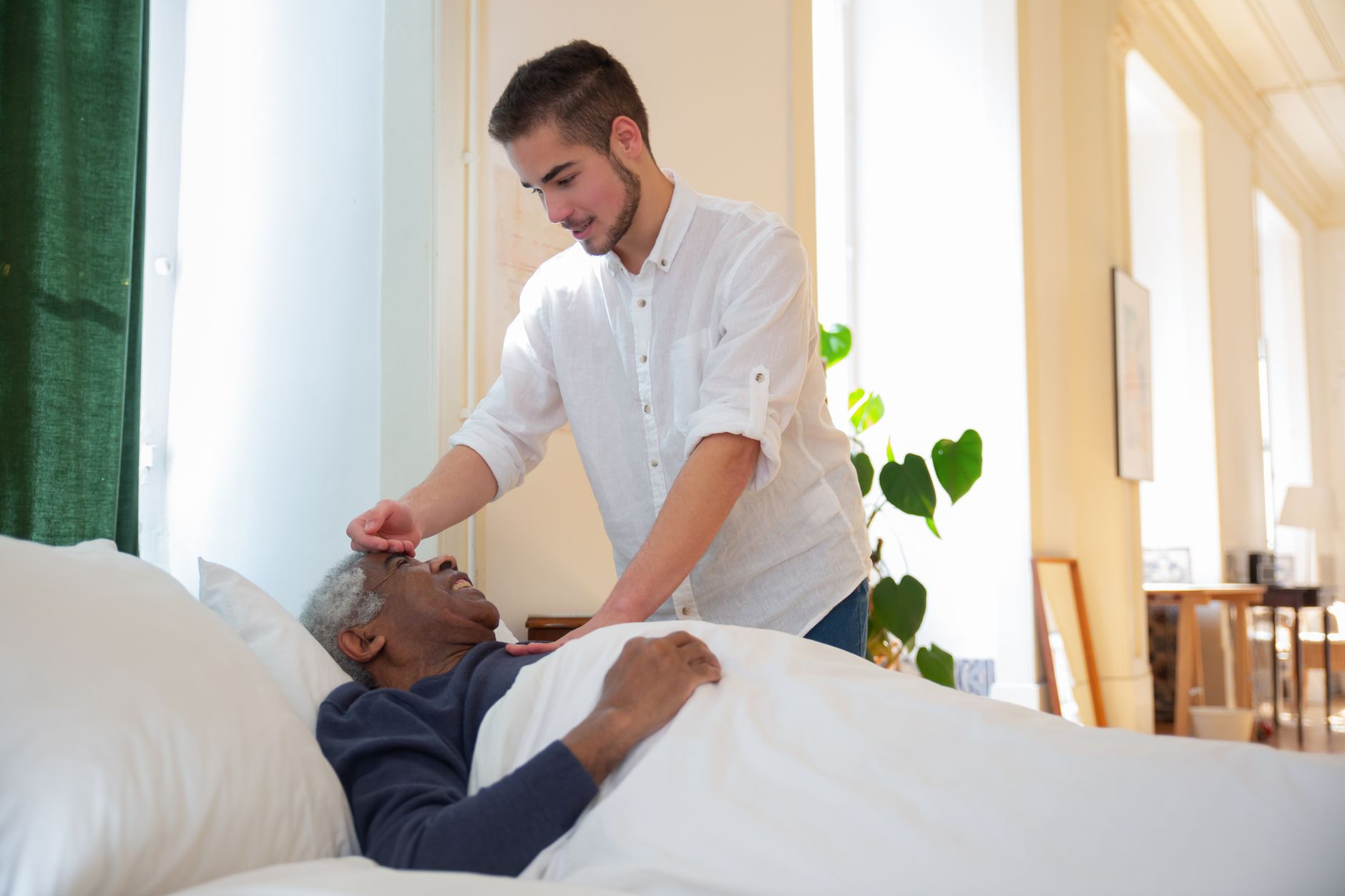 Nệm tốt nhất cho người già trên 60 tuổi - A young man supports an elderly man in bed, offering care in a bright room.