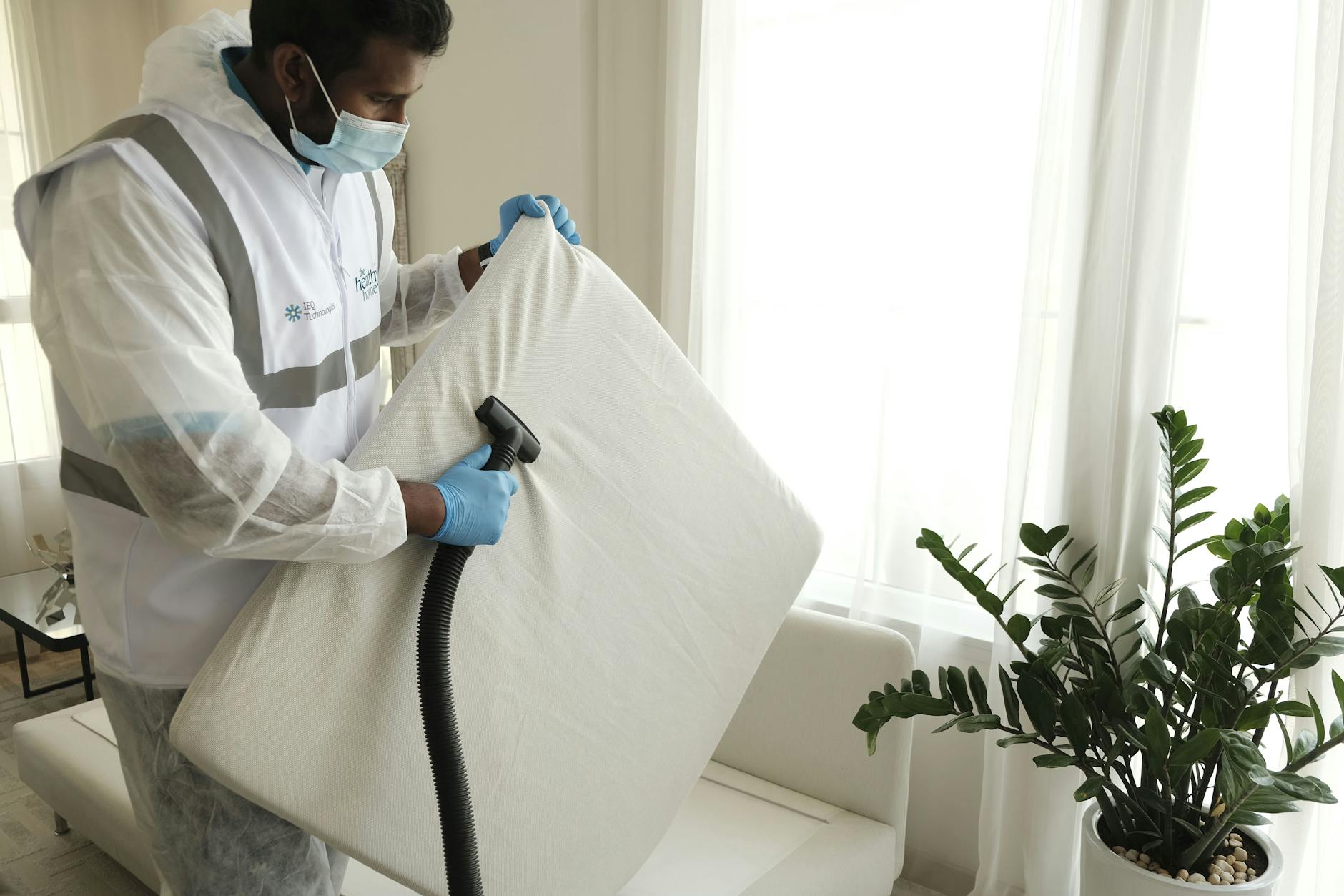 Xử lý nệm bị mốc, hôi, xẹp lún. - Man in protective gear vacuuming a mattress in a modern home interior.