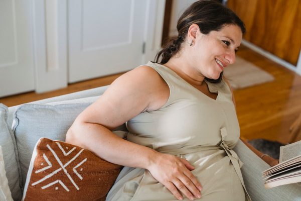 Nệm phù hợp cho bà bầu 3 tháng cuối - Young pregnant female in beige dress smiling and looking away while reading book on comfortable sofa