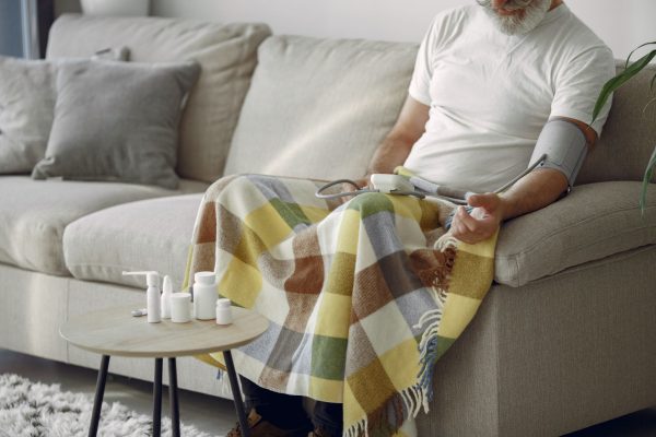 Nệm tốt cho người cao huyết áp - Elderly man monitoring blood pressure on a cozy couch at home, surrounded by medication.