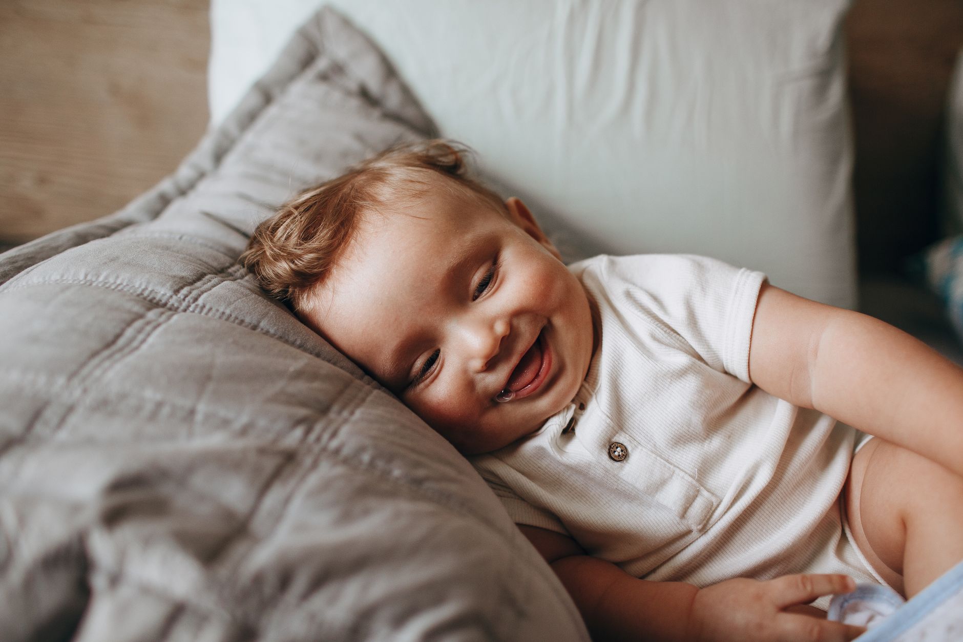 Nệm cho bé từ 1 đến 3 tuổi loại nào tốt - A cute baby in a white romper smiles happily while lying on a soft pillow.