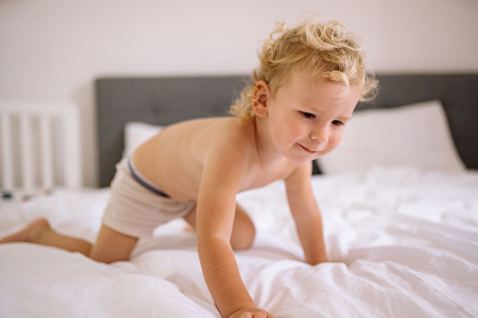 Nệm cho bé từ 1 đến 3 tuổi loại nào tốt - A playful toddler with curly hair is crawling on a bed in a well-lit home bedroom setting.